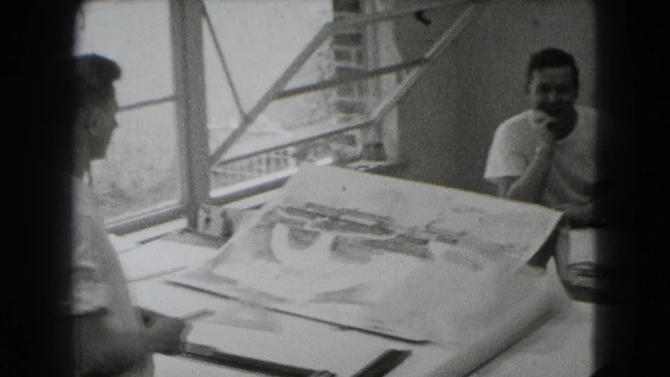 Three men appear in a black-and-white Middletown interior, with large plans spread across a table by an open window.