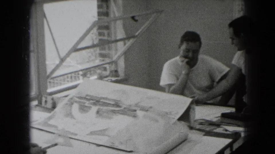 Black-and-white 8mm frame showing a seated man at a plan-covered table, with another figure partly visible nearby.
