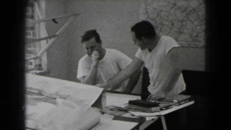 Two men in a black-and-white Middletown home-movie frame lean over large plans in an informal workspace.