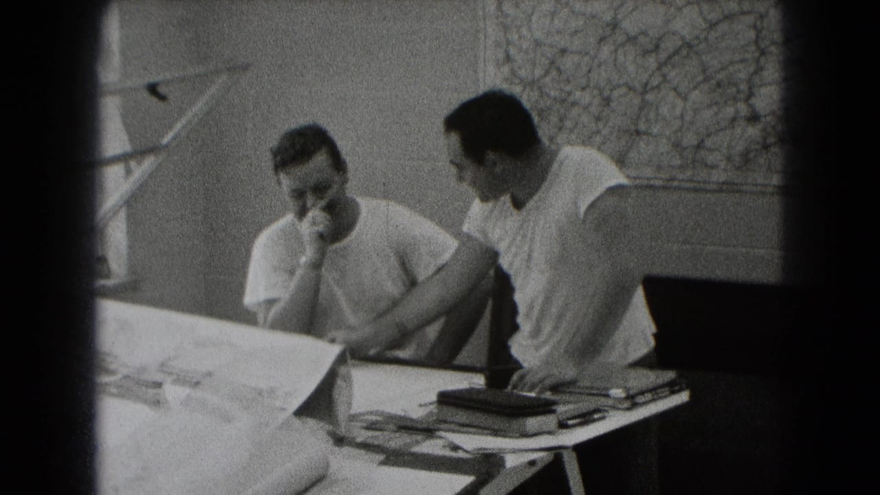 Two men in white T-shirts sit at a table with papers or plans in an interior work area.
