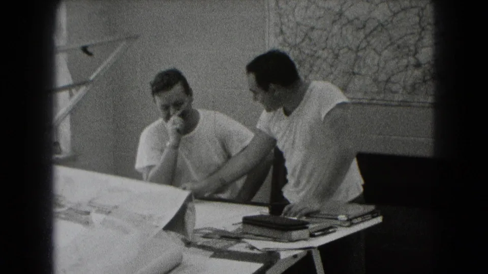 In Middletown, Pennsylvania archival 8mm footage, two adult men sit by a drafting table with papers, tools, and a wall map behind them.