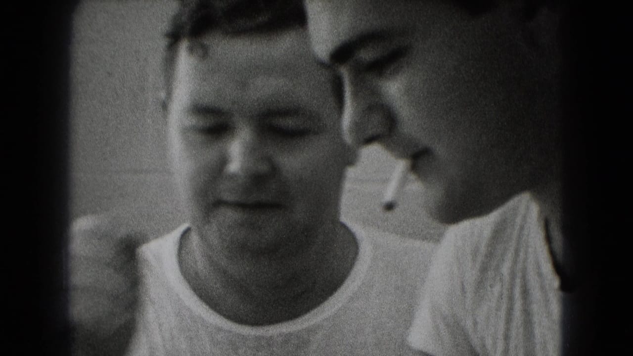 Close 8mm frame of two adult men in a Middletown work interior, one smoking while the other looks down toward plans.