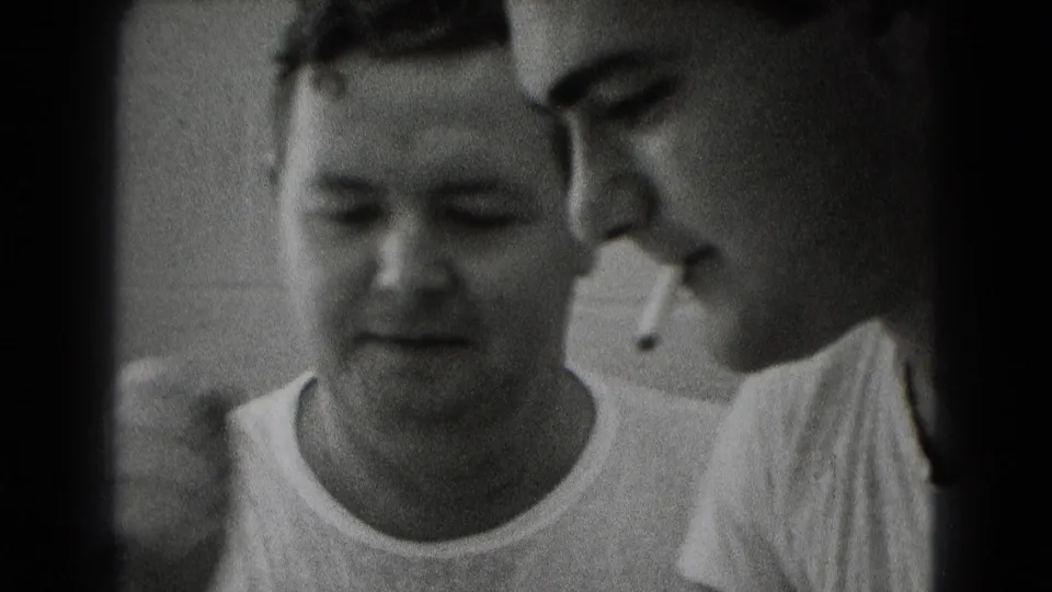 Close 8mm frame of two adult men in a Middletown work interior, one smoking while the other looks down toward plans.