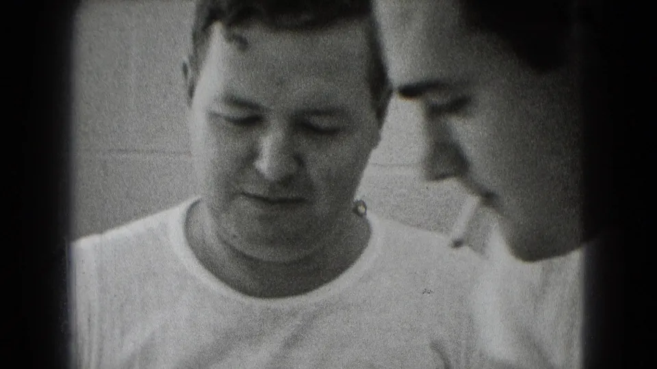 Close archival frame of two men in a private Middletown work area, with a cigarette visible as they focus on something below frame.