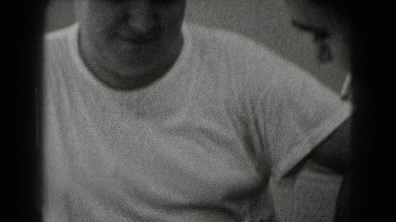 Seated man in a white T-shirt seen close-up in a grainy black-and-white frame.