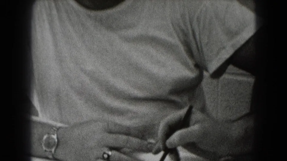Grainy black-and-white frame of an adult man seated indoors while another person reaches toward his forehead in a playful gesture.