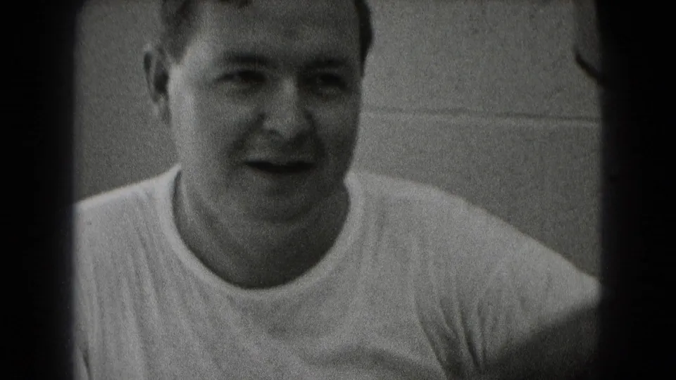 Close black-and-white 8mm frame of a smiling adult man in a white T-shirt seated indoors, with another person partly visible at right.