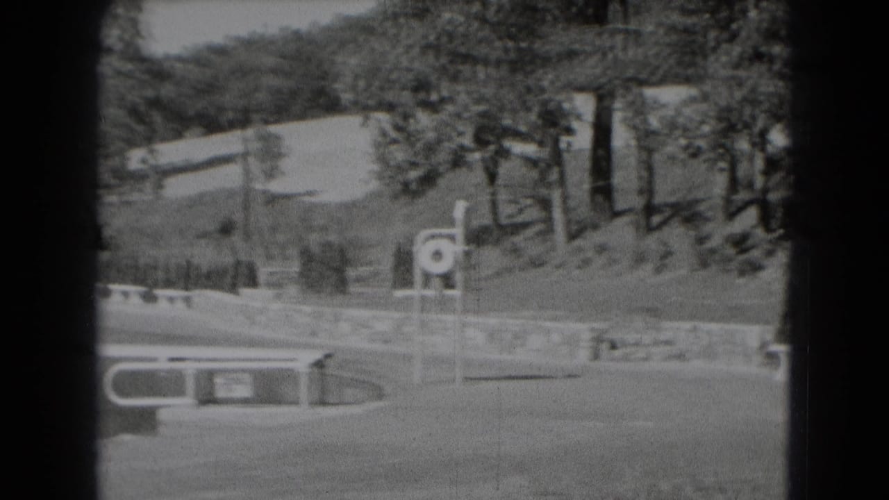 Open field with trees, low wall, bench, and circular signpost beyond water.