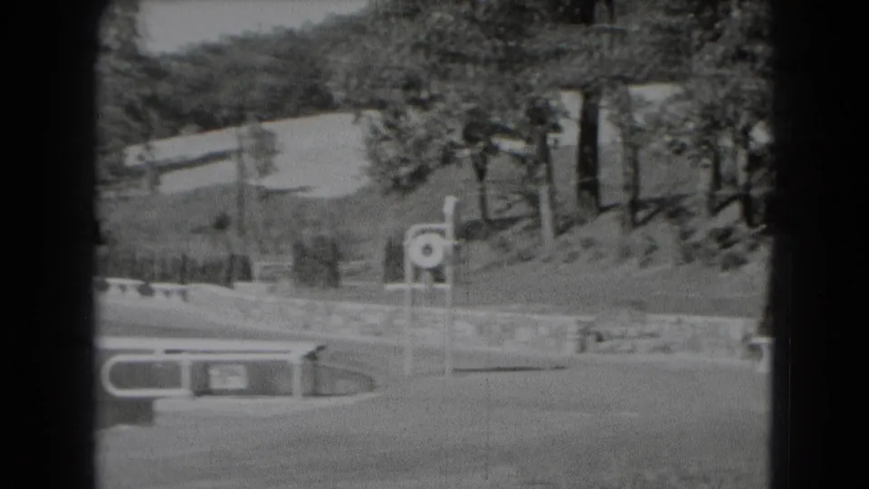 1975 8mm view in Middletown, Pennsylvania, showing open grounds, trees, a low wall, a bench, and a circular signpost beyond water.