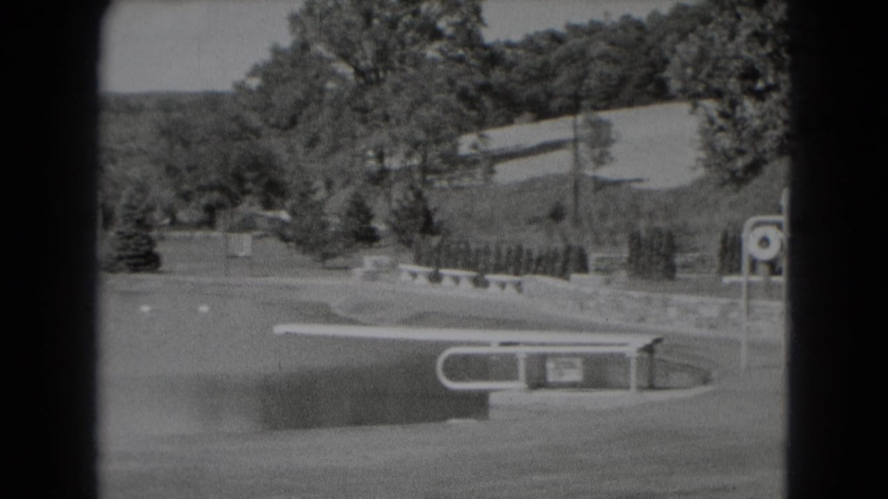 Middletown, Pennsylvania home-movie frame from 1975 showing a landscaped field edge with trees, a low wall, bench, and signpost near water.