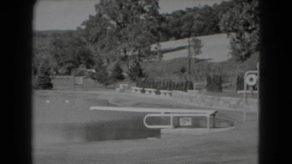 Middletown, Pennsylvania home-movie frame from 1975 showing a landscaped field edge with trees, a low wall, bench, and signpost near water.