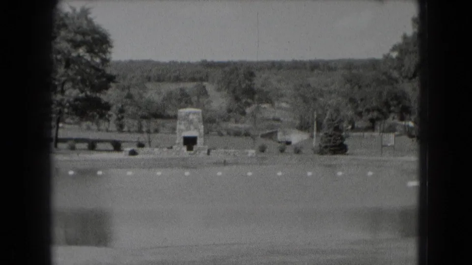 Black-and-white 8mm frame from 1975 Middletown, Pennsylvania, showing water, open grounds, a low wall, benches, trees, and a stone structure.