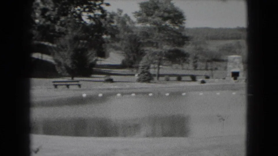 Black-and-white 8mm view of a Middletown park pond with buoys, a bench, trees, lawn, and a stone marker in the background.