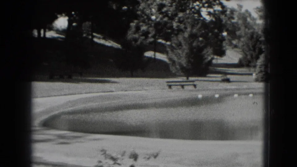 1975 8mm park footage shows a curved pond edge with buoys, a bench, trees, and shaded grass in Middletown, Pennsylvania.
