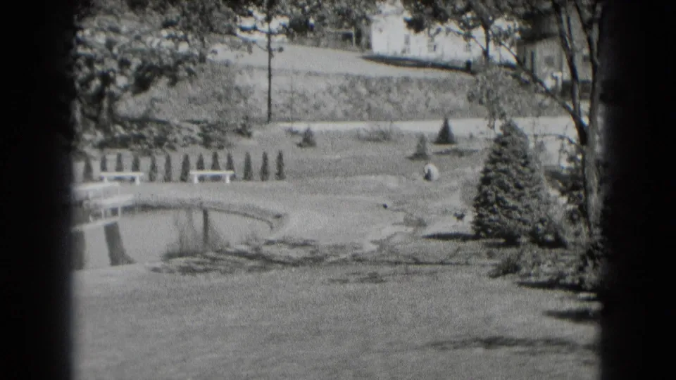 Black-and-white 8mm frame of a Middletown park landscape with pond edge, open lawn, shrubs, evergreen tree, and sloped background.