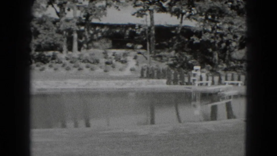 1975 home-movie frame looking across still water toward benches, landscaped shrubs, mature trees, and a shaded structure or crossing.