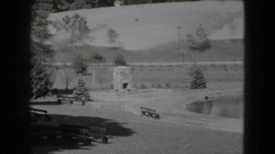 A late frame shows park benches in shade, a stone outdoor fireplace, small evergreens, curved lagoon edge, and open hillside in the background.