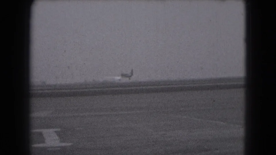 Small propeller aircraft near the runway horizon, with foreground pavement markings in 1975 Middletown home-movie footage.