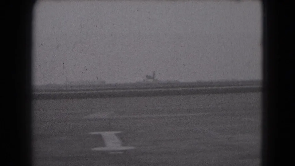 Distant small aircraft on a Middletown runway, with visible pavement marking and grainy 8mm film texture.
