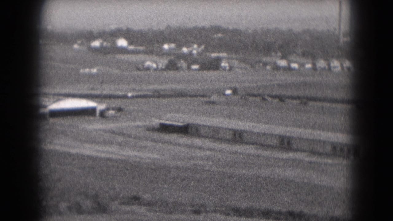 1975 Middletown, Pennsylvania 8mm frame showing a long rectangular building, moving vehicles, open ground, and distant smokestacks.