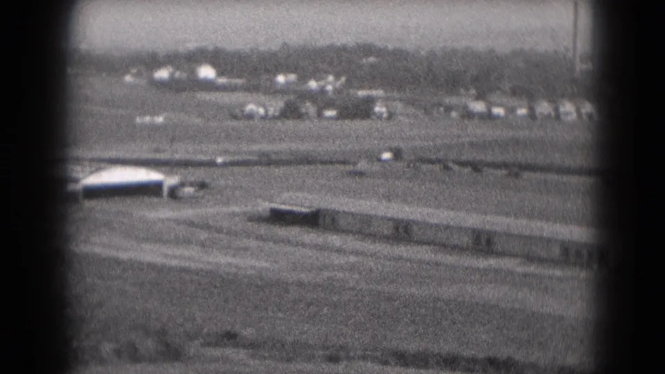 1975 Middletown, Pennsylvania 8mm frame showing a long rectangular building, moving vehicles, open ground, and distant smokestacks.