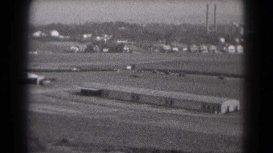 1975 Middletown, Pennsylvania view of vehicles moving behind a long low structure, with trees and factory stacks in the distance.
