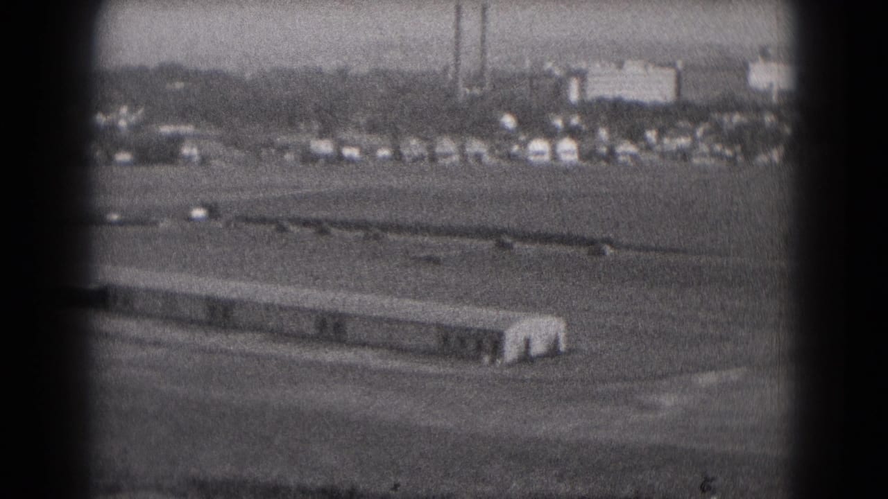 1975 Middletown, Pennsylvania 8mm frame showing a distant aircraft on an open airfield with cars, buildings, and smokestacks in the background.
