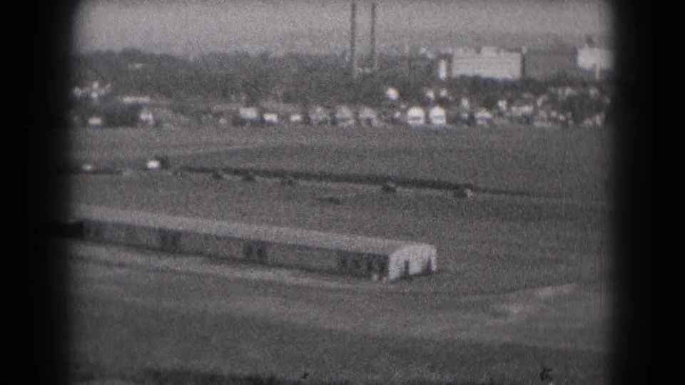 1975 Middletown, Pennsylvania 8mm frame showing a distant aircraft on an open airfield with cars, buildings, and smokestacks in the background.