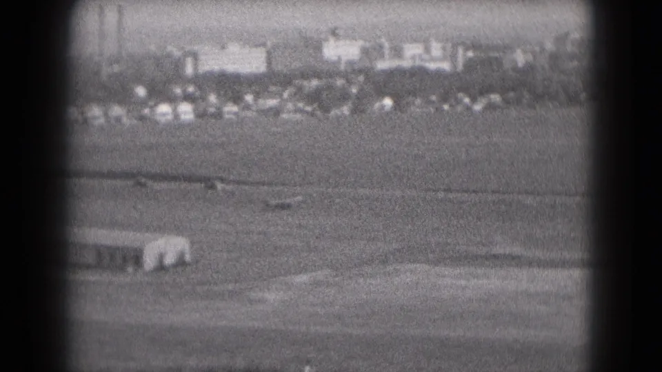 1975 8mm airfield view in Middletown, Pennsylvania, showing a distant small aircraft below a hazy line of cars, buildings, and stacks.