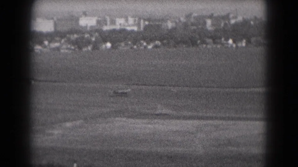 Final 1975 Middletown airfield frame with a small aircraft on the open runway area, backed by parked cars, low buildings, and distant stacks.