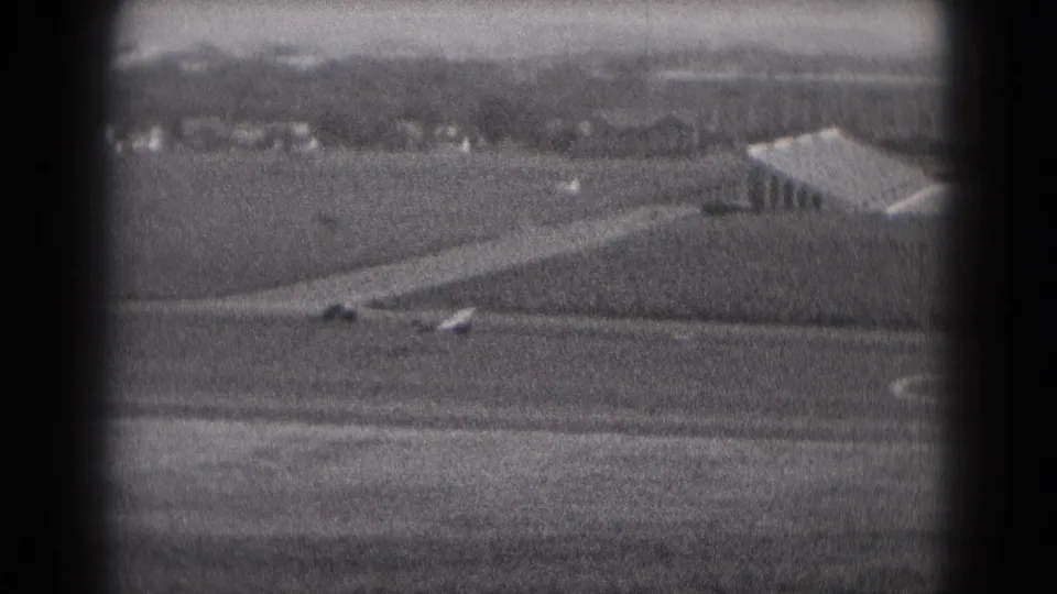 A distant small plane passes a rural runway area with a low building visible in 1975 black-and-white 8mm footage.