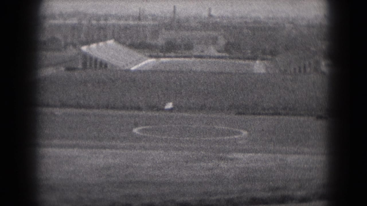 Small airplane on or near a runway with open fields and distant buildings.