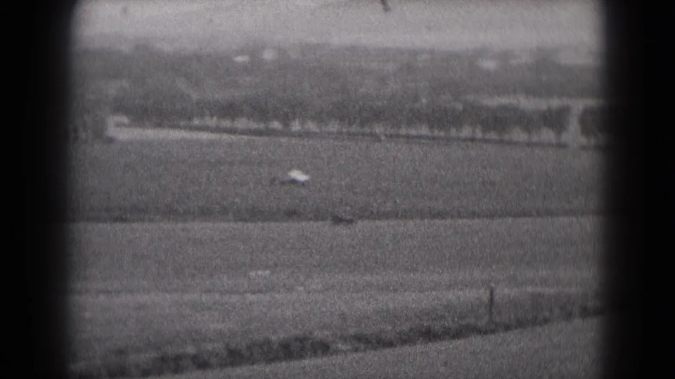 Small aircraft appears distant over the Middletown airfield landscape, with 1975 black-and-white 8mm grain and soft focus.