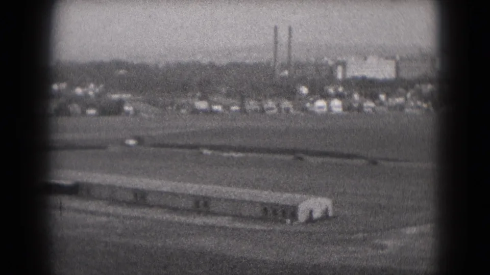1975 home-movie frame of airport-edge grounds near Middletown, with a low building, open field, vehicles, tanks, and smokestacks.