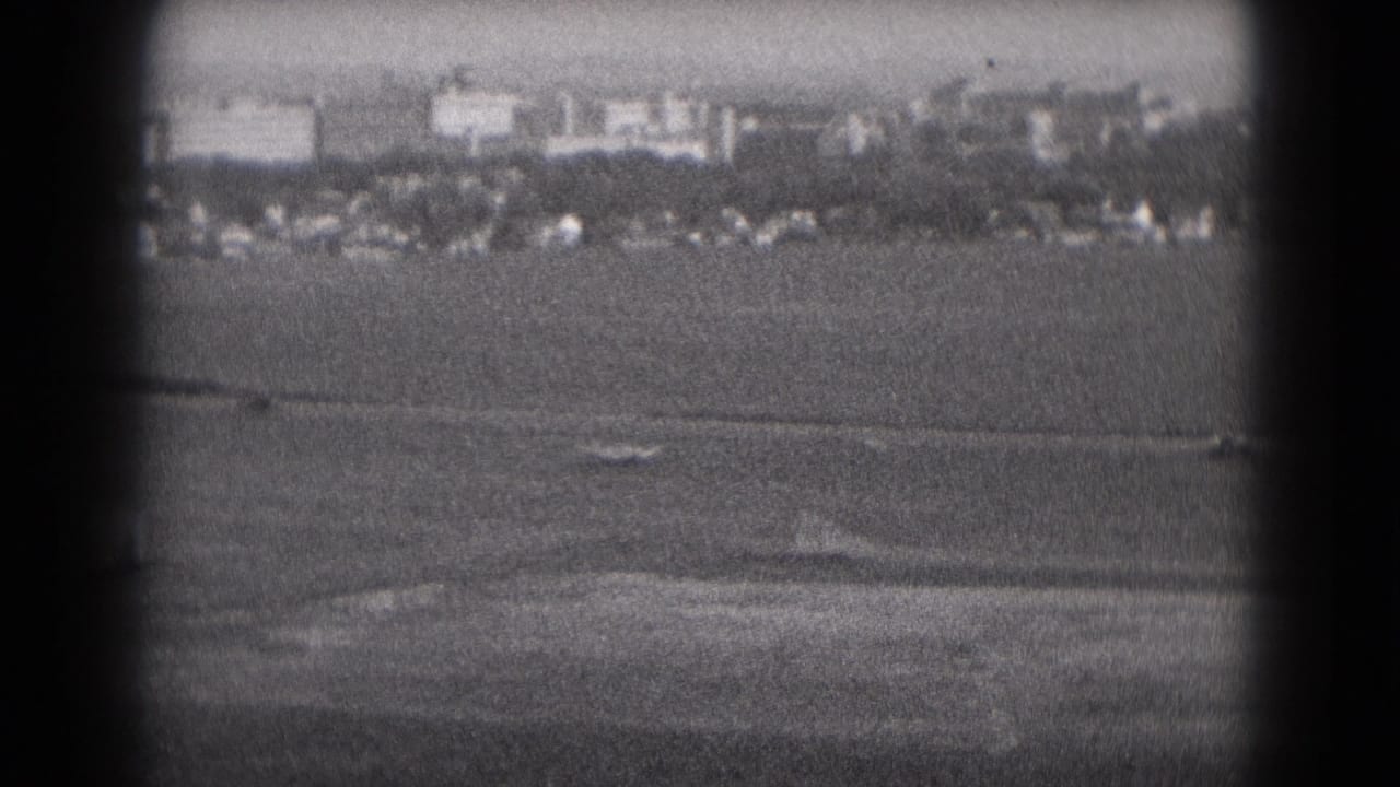 A soft black-and-white 8mm frame shows open land and distant Middletown-area buildings partly obscured by the camera gate shadow.
