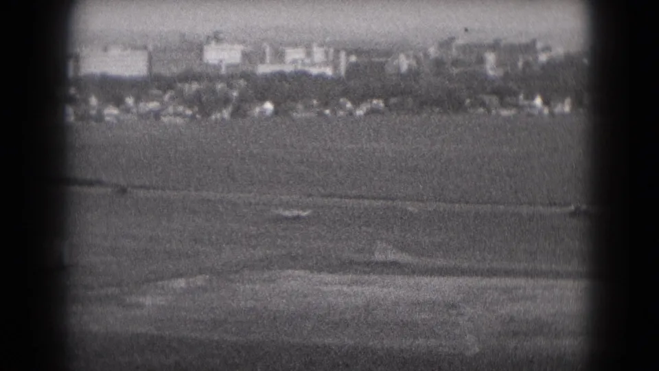 A soft black-and-white 8mm frame shows open land and distant Middletown-area buildings partly obscured by the camera gate shadow.