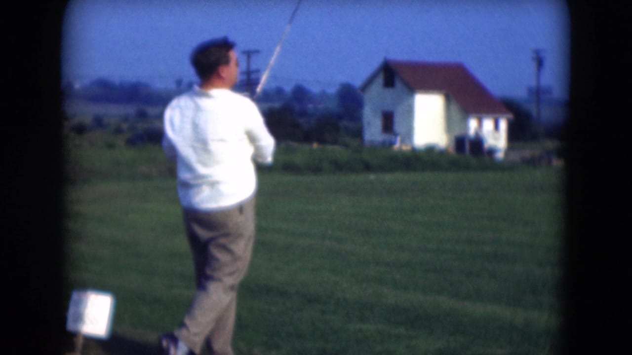 Man in a white shirt swings a golf club on a grassy driving range.