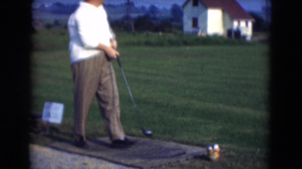 A man at a Middletown, Pennsylvania driving range stands with a golf club on a tee mat near a bucket of balls in 1975 home-movie footage.