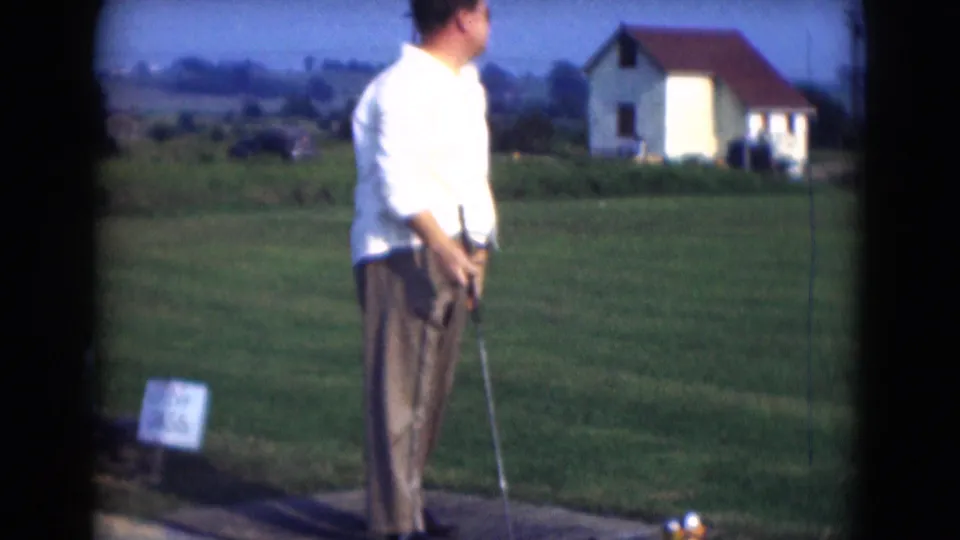 A man in a white shirt and brown trousers prepares at a driving-range tee in 1975 Middletown, Pennsylvania home-movie footage.