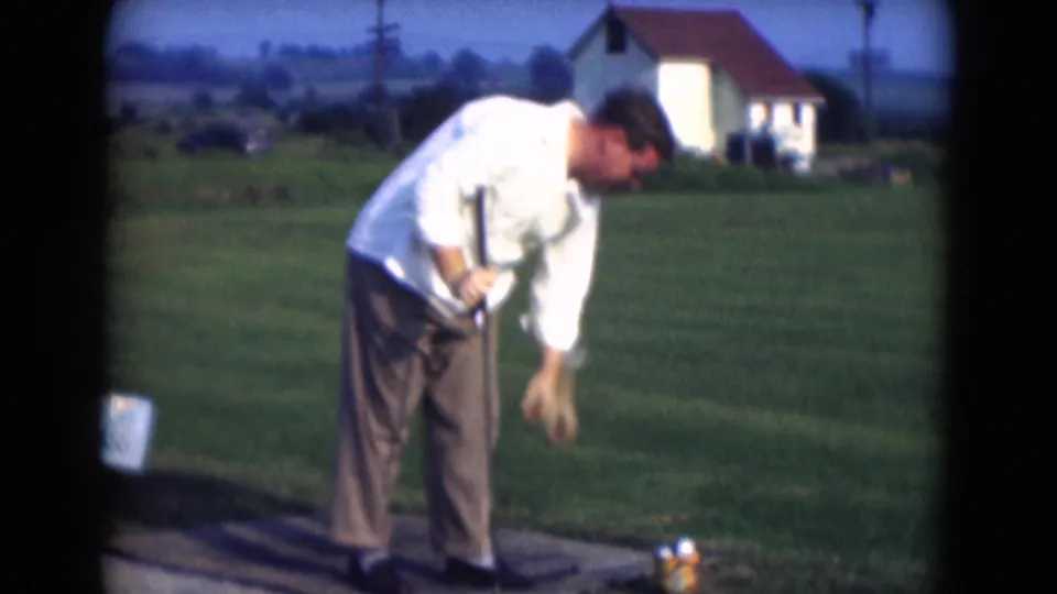 The golfer bends near the tee area and ball bucket during a 1975 Middletown, Pennsylvania driving-range home movie.