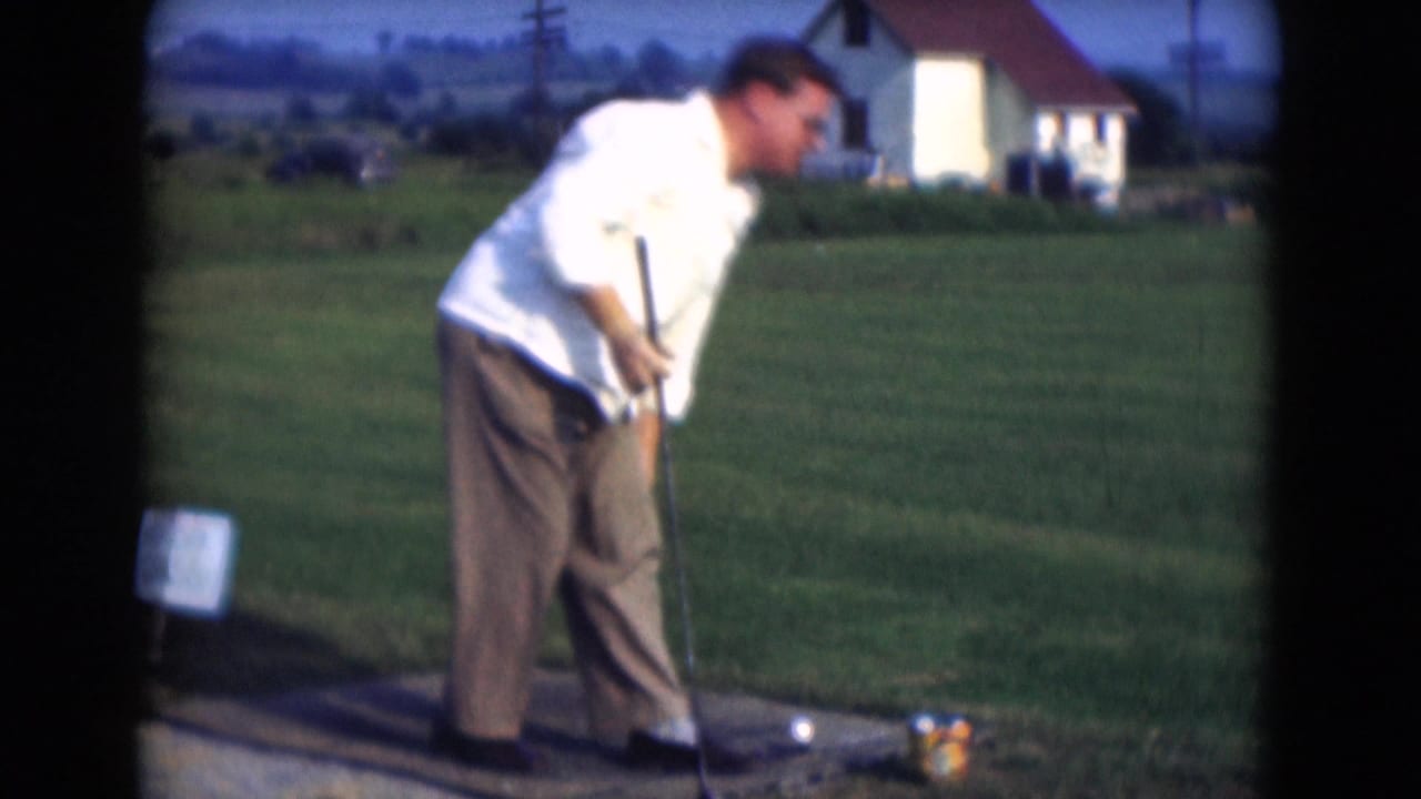 A man in a white shirt addresses a golf ball with a club on a grassy field in 1975 Middletown, Pennsylvania.