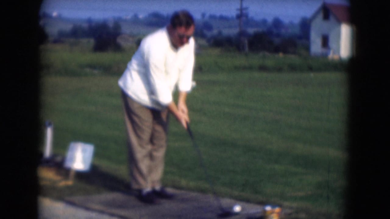 In Middletown, Pennsylvania in 1975, a man in a white shirt sets up a golf swing on an outdoor practice mat near grass and a small building.