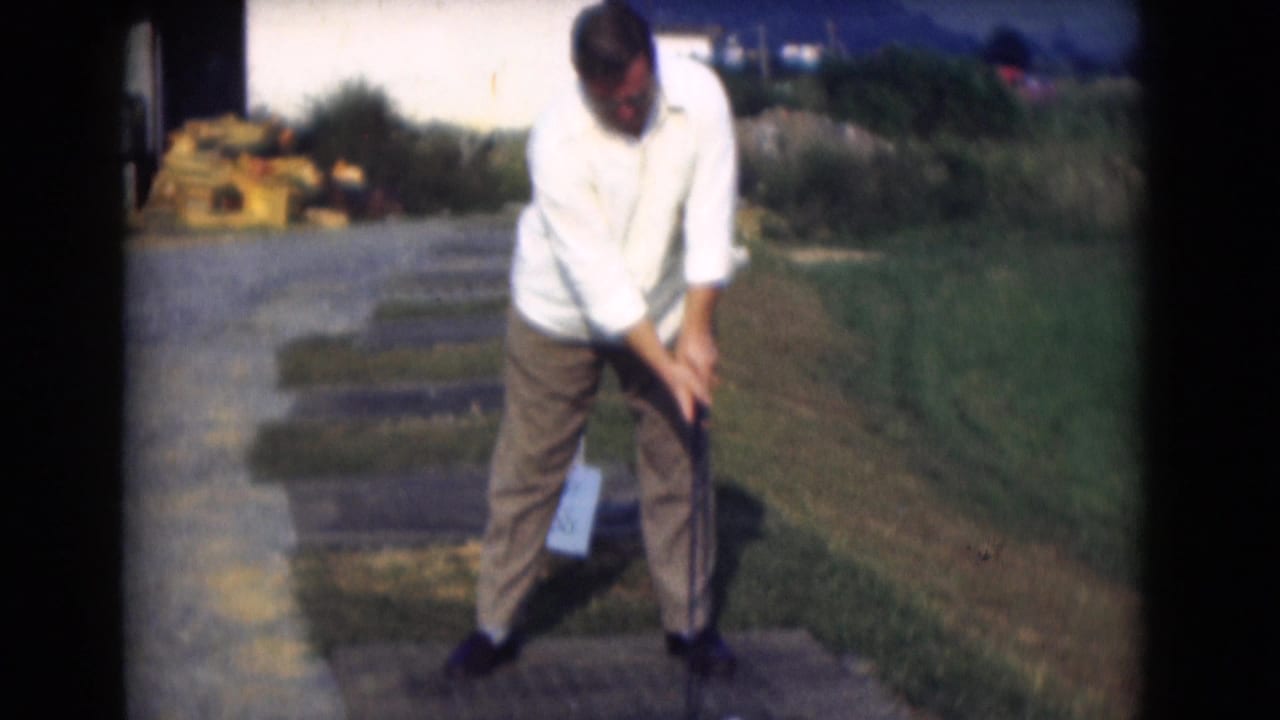 The golfer stands over the ball in a 1975 Middletown, Pennsylvania home-movie frame, with a paved practice area and grass behind him.