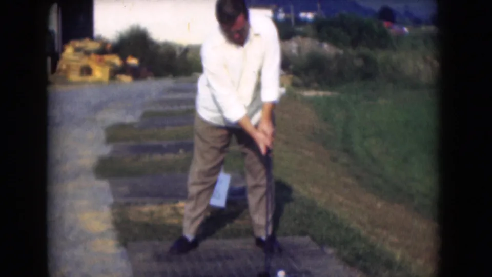 A man in a white shirt and brown trousers holds his golf club low over the ball at an outdoor practice area in 1975 Pennsylvania footage.