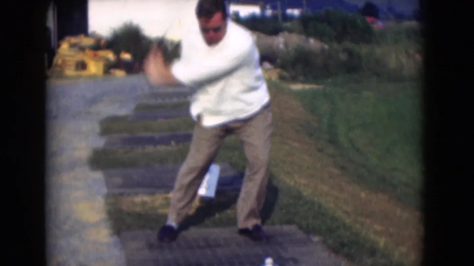 In the closing frame, the golfer turns through his swing on the outdoor mat, with grass, pavement, and distant buildings visible around him.