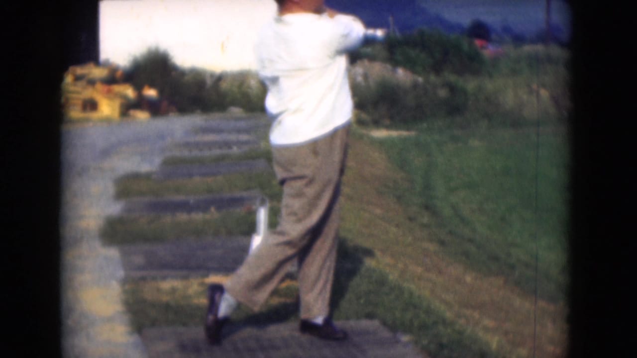 In 1975 Middletown, Pennsylvania, an adult man in a white shirt and brown trousers follows through on a golf swing near grass and paving.
