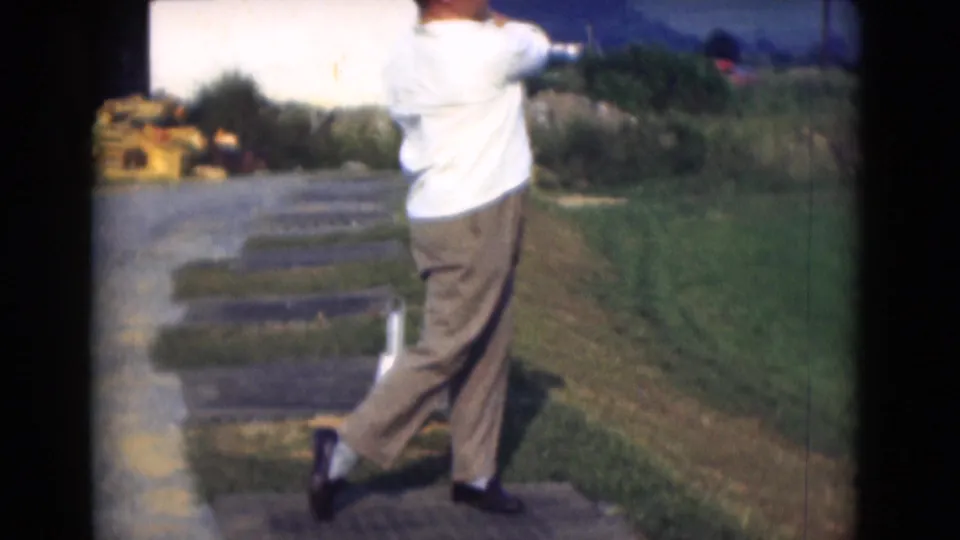 In 1975 Middletown, Pennsylvania, an adult man in a white shirt and brown trousers follows through on a golf swing near grass and paving.