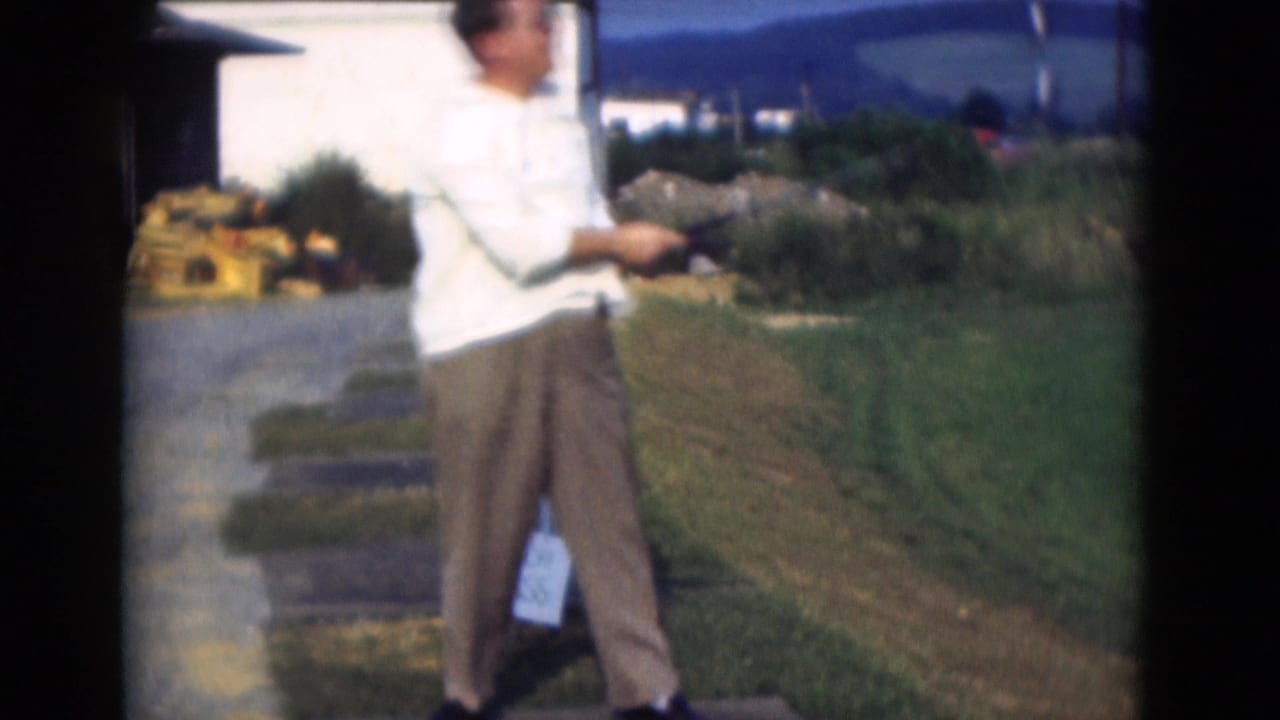 The golfer turns after his swing in Middletown, Pennsylvania, with a paved practice strip, grass, low buildings, and hills in the background.