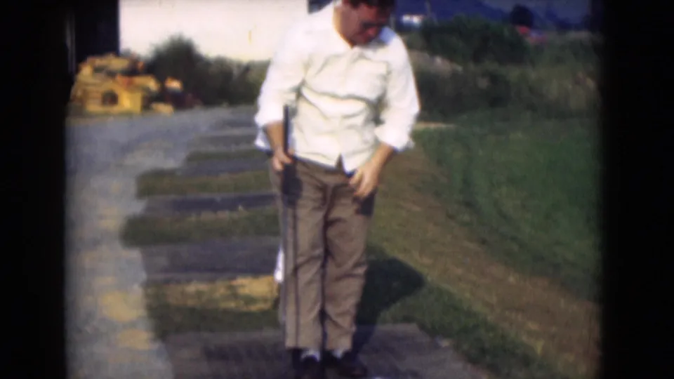 An adult golfer stands between swings on a sunny 1975 Middletown practice area, with grass, paving, and soft 8mm color visible.