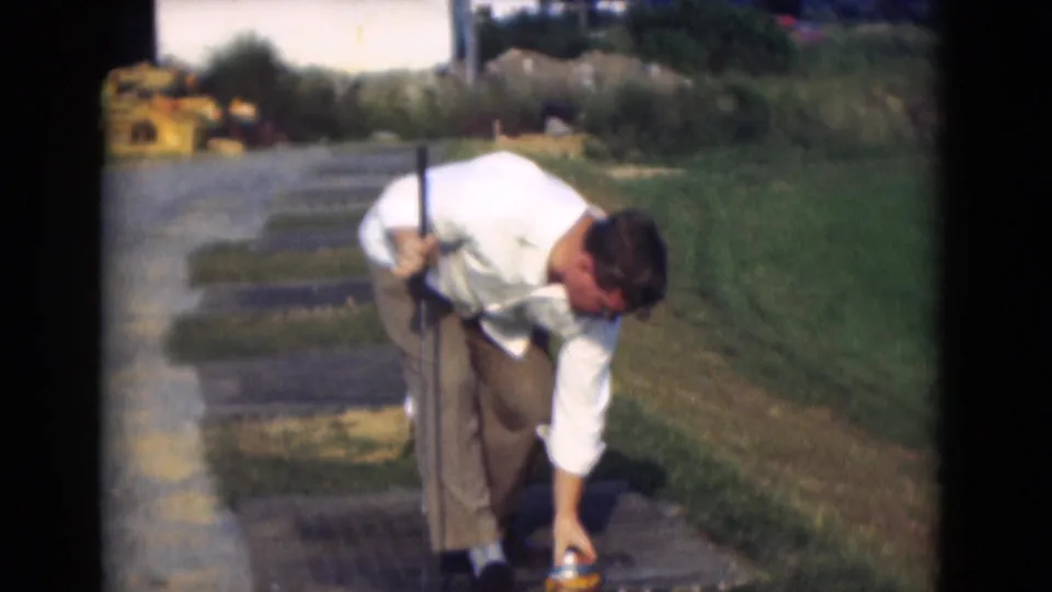 The golfer bends to reach toward the tee or ball area while holding his club, with grass, paving, and bright 8mm exposure around him.